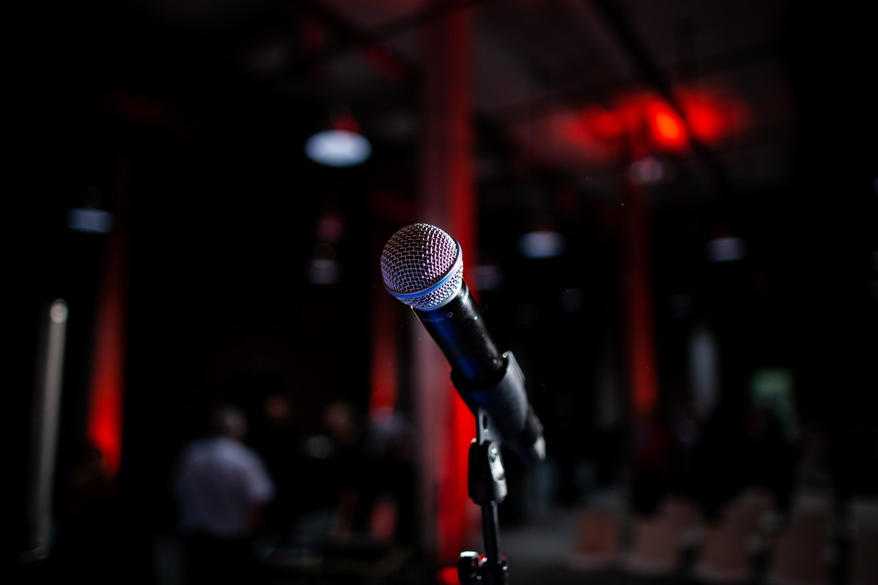 Microphone is seen at close range, on a stand. In the background, out of focus is seating.