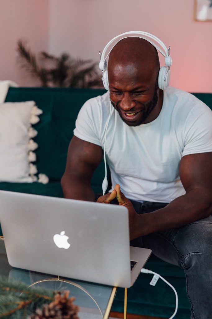 Man sits on couch with over the ear headphones on while looking at an Apple laptop on lowered table in front of him